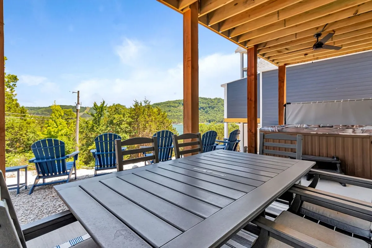 A spacious wooden deck featuring a large outdoor dining table, blue Adirondack chairs, and a private hot tub, all set against a lush green landscape. The arrangement invites both dining and relaxation while offering panoramic natural views, and the multi-level design hints at expansive, versatile outdoor spaces.