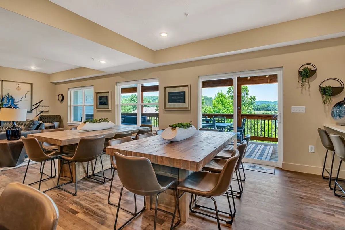 This image features a large communal dining table set in a welcoming living/dining space with comfortable seating, large windows, and warm lighting. The windows provide views of the outdoors, and the arrangement encourages both shared meals and relaxed conversation. Plush seating and tasteful decor create an inviting environment for group gatherings.