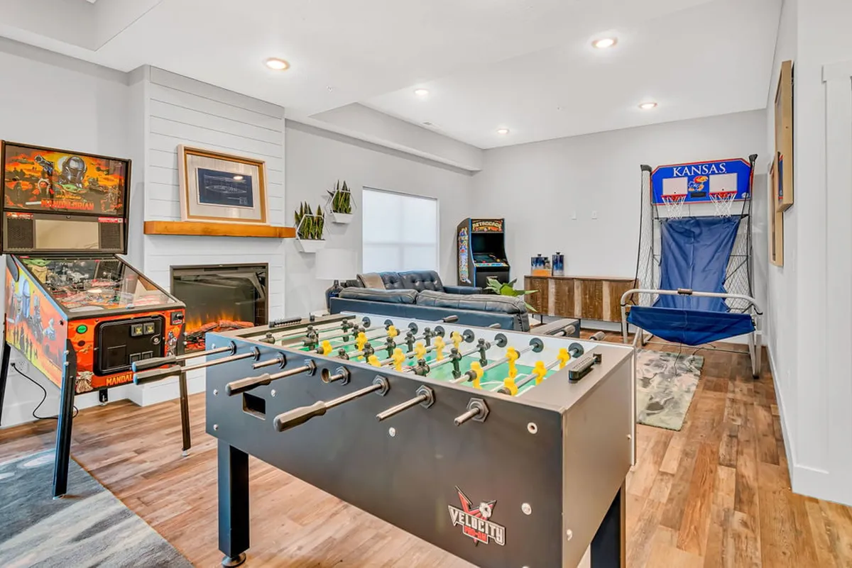 The image showcases a vibrant game room featuring a modern foosball table, classic pinball machine, retro arcade game, a plush sectional sofa, and a cozy fireplace. The scene suggests a lively, welcoming space perfect for group entertainment and relaxation.