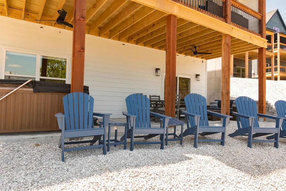 This image captures a welcoming wooden deck with blue Adirondack chairs against a white exterior wall, inviting guests to relax outdoors. The setting is casual and inviting, with plenty of space that suggests a comfortable spot for guests to enjoy the fresh air—perfect for lounging with a small dog by your side.