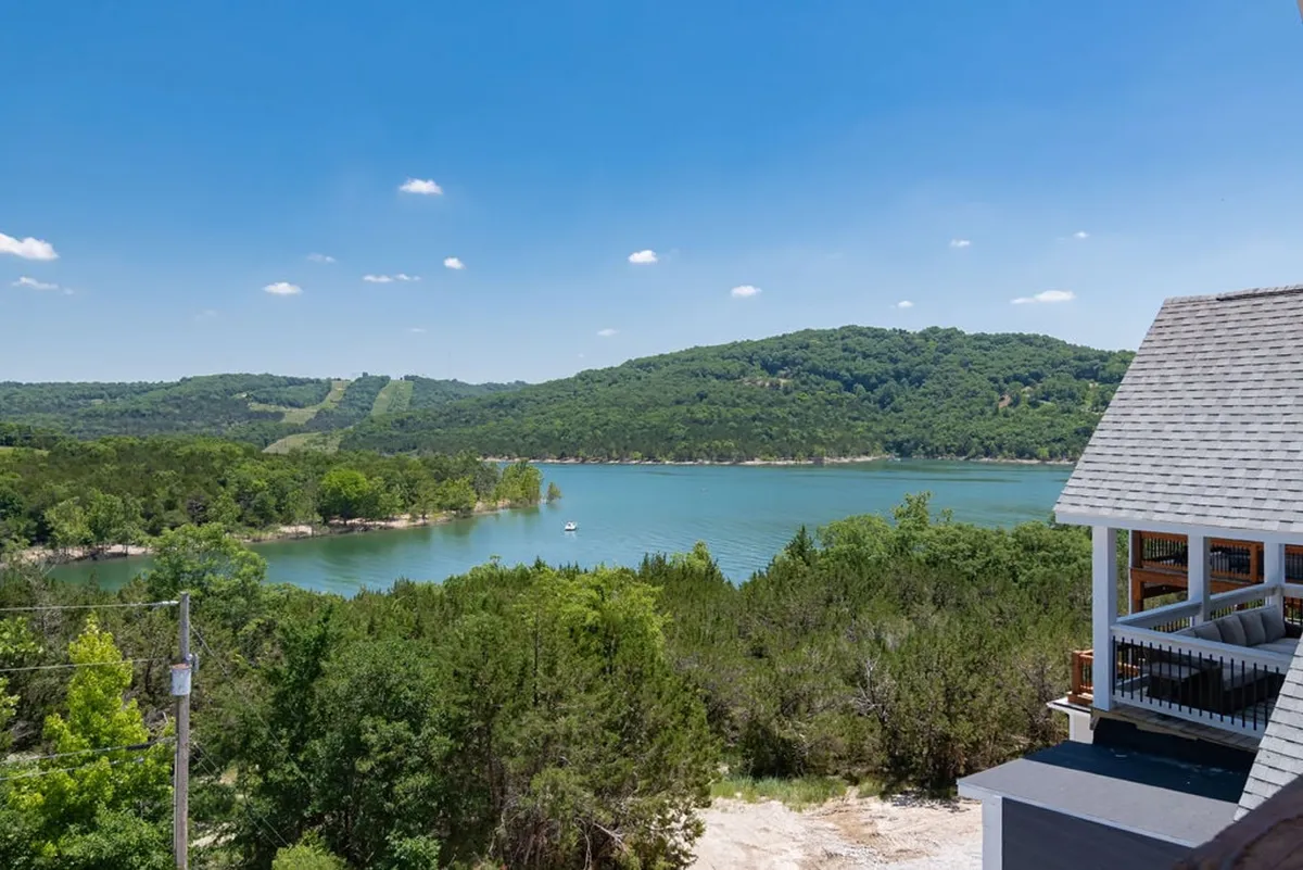 A tranquil view of Table Rock Lake, with rolling hills in the background and a house balcony in the foreground. The lake's calm waters are framed by nature, suggesting immediate access to swimming, fishing, kayaking, or simply relaxing and soaking in the scenery. The vantage point from the balcony connects the private home to the lake experience.