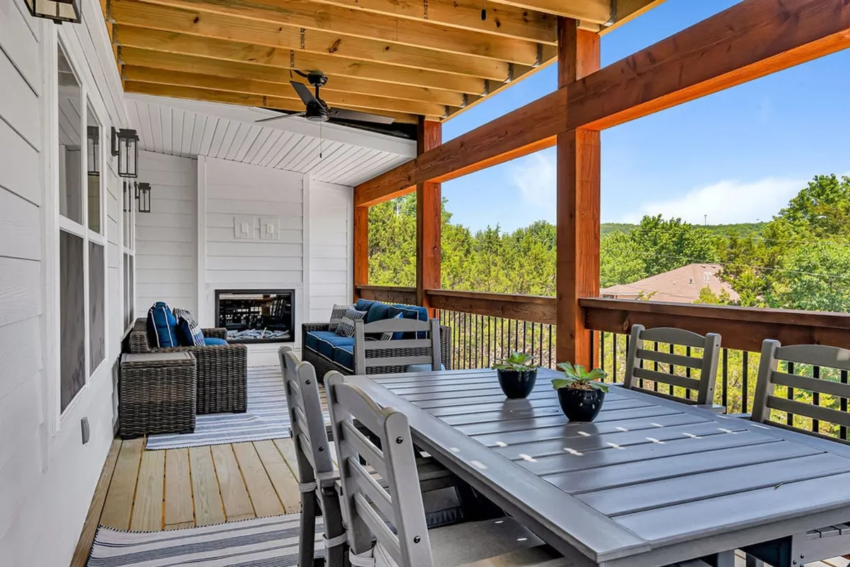 A comfortable seating area and dining table are arranged on an outdoor patio, with a stone fireplace as the centerpiece and abundant greenery in the backdrop. The space is set for group gatherings, with soft lighting suggesting a welcoming ambiance for day or night.