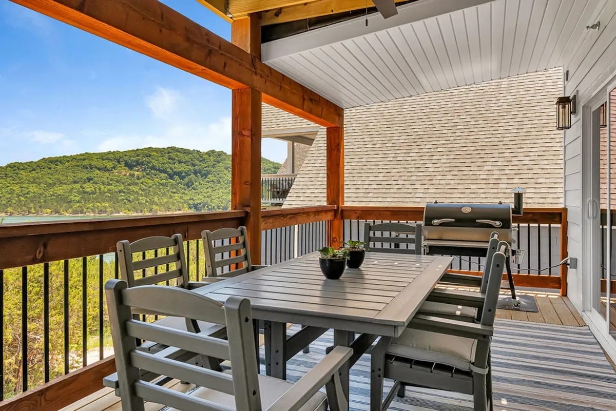An exterior scene with a large outdoor dining table next to a high-end grill, set on a patio overlooking a scenic hillside. The setting is ideal for evening barbecues or casual get-togethers, with a backdrop that hints at festive and relaxed occasions.