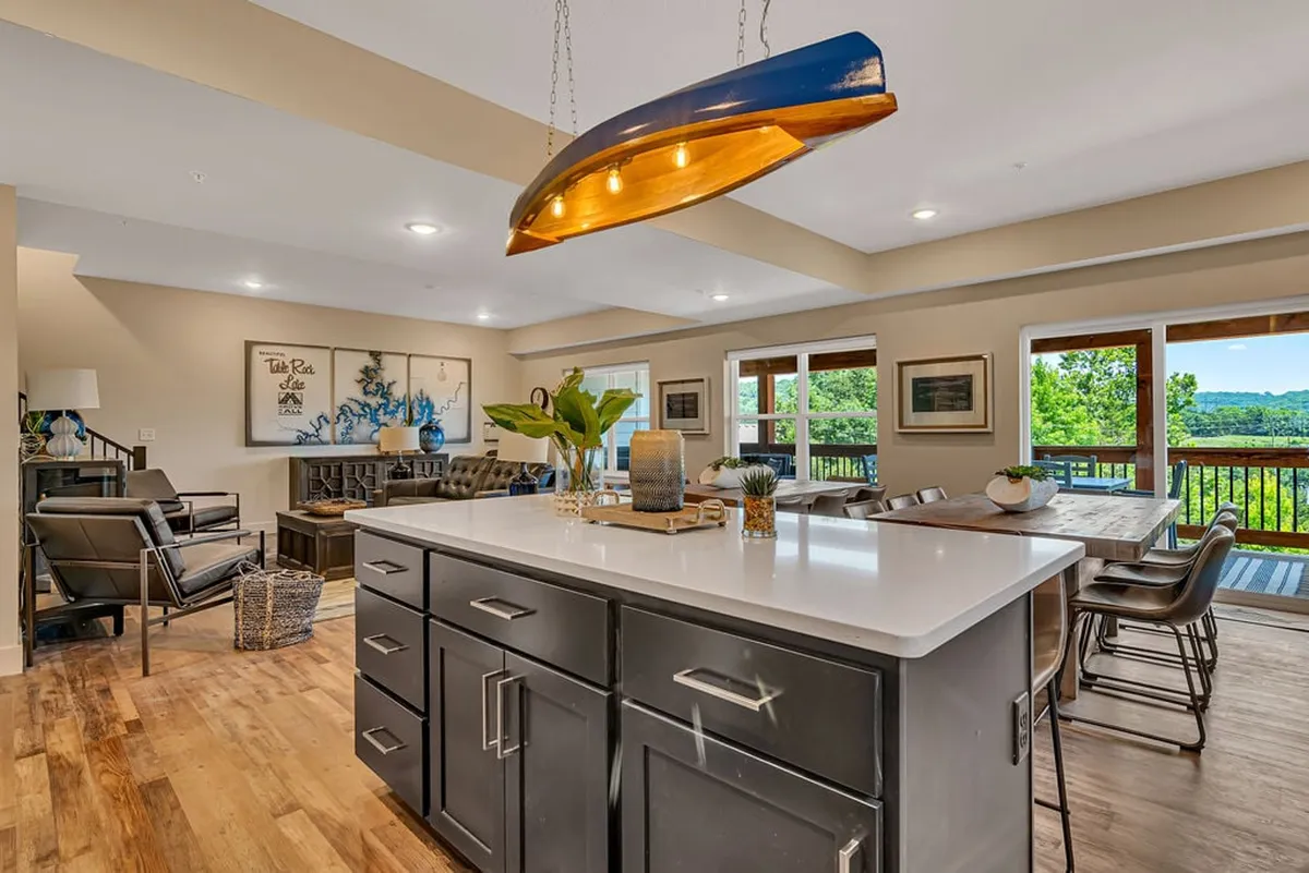 A bright, welcoming kitchen and dining area with a central island, hanging light fixture, and a large window overlooking scenic views. The space combines modern comforts with warmth, featuring a spacious kitchen ideal for preparing family recipes and an adjacent dining area perfect for celebrations or casual family dinners.