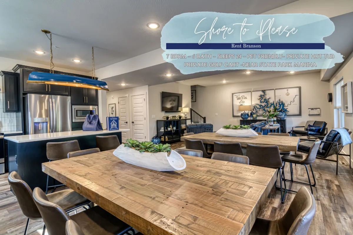 This image showcases a spacious, open-concept living and dining area that includes a large wooden dining table, a stylish blue pendant light, and a comfortable lounge space. The adjoining modern kitchen features sleek appliances, and large windows flood the room with natural light, revealing views of the surrounding landscape. The overall ambiance is inviting and designed for communal gatherings.