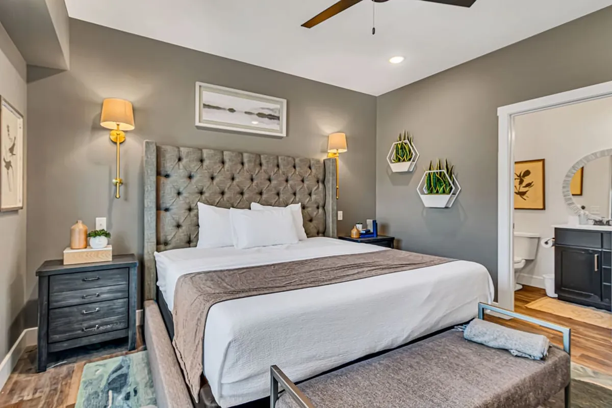 A serene bedroom featuring a tufted headboard, crisp white linens, nightstands with lamps, wall art, and hanging planters, all softly lit by natural light streaming through a nearby window. The atmosphere radiates tranquility and comfort, ideal for relaxation and unwinding after a busy day.
