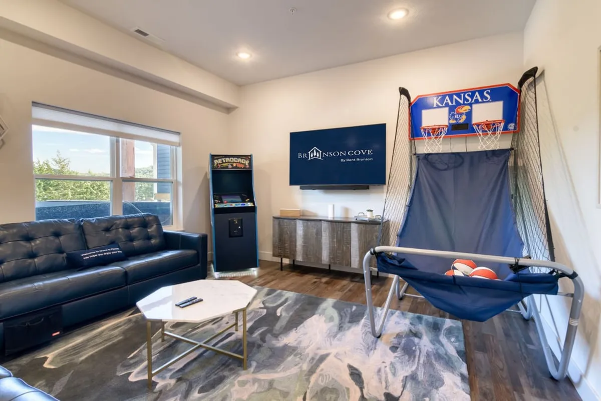 A brightly lit game room featuring a sofa, arcade machine, basketball hoop, and coffee table. The space is casual and fun, suggesting indoor entertainment options for families or groups to enjoy together during downtime or rainy days.